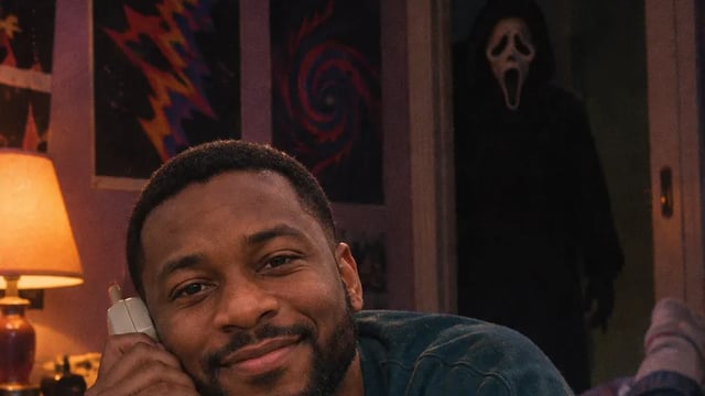 Man on a satin bed talking on a corded phone while Ghostface watches from the doorway
