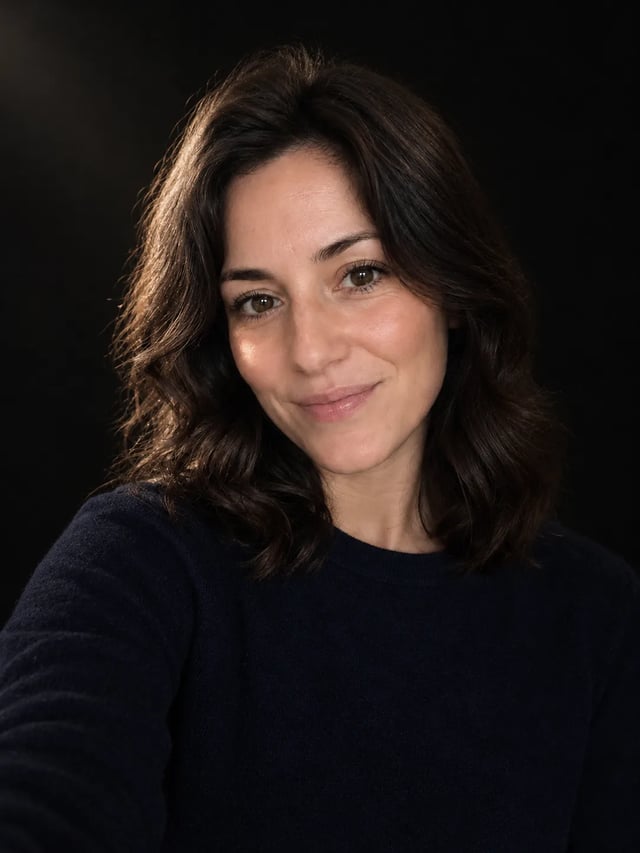 The woman from the kitchen selfie on a near-black backdrop, lit by one soft light from the left of the frame, with a warm glow on her cheek and hair