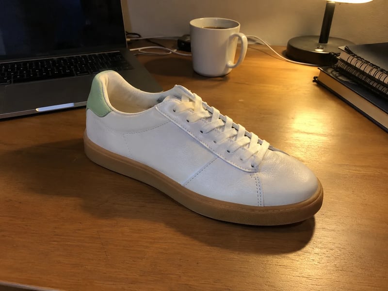 White leather sneaker with a gum sole on a wooden desk under a yellow lamp, next to a laptop and a coffee mug